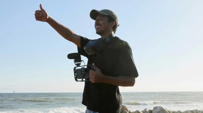 Image of filmmaker shooting showing a thumbs up with an impressive set of equipment mounted to their phone. Standing at the beach.