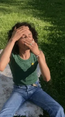 A person sits on the grass on a blanket with dark curly hair and tan skin and rubs oil on their skin