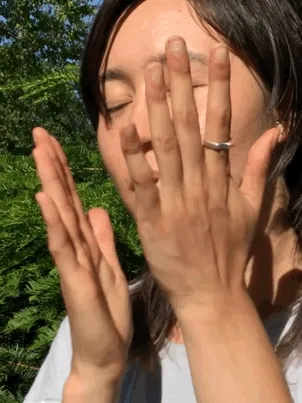 An Asian person with shaggy hair presses oil onto their faces and smiles sweetly at the camera