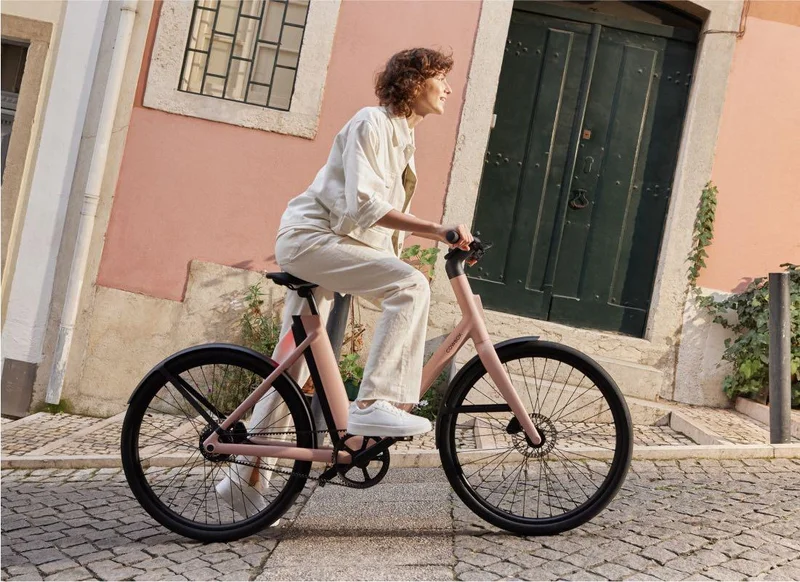 A photograph. A woman sits on a Cowboy Cruiser ST and prepares to push off. The street is apparently a steep hill but the photograph has been composed so that the cyclist appears to be on a flat incline..