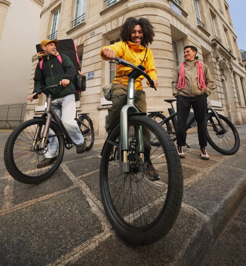 A group of three cyclists pause on their Cowboy bikes in a city setting, their expressions relaxed and joyful. The bikes, in various colourways, are parked on stone pavement near a historic European-style building with arched windows and beige walls.