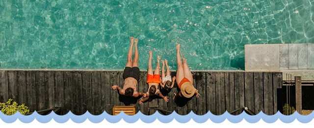 Top view of four people relaxing on a wooden deck beside a clear blue swimming pool.