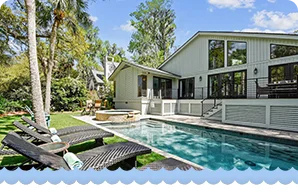Modern home with large windows, a sleek pool, and lounge chairs surrounded by lush greenery.