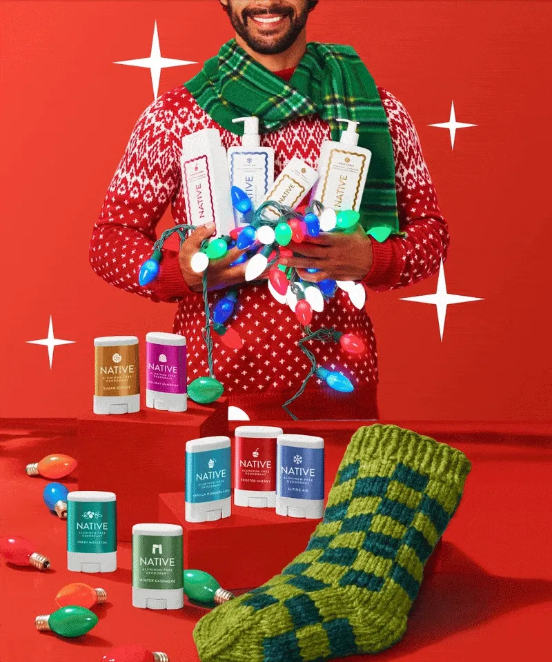 Man wearing festive sweater holding Native products, a table of mini deodorants sits in front of him. Man wearing festive sweater holding Native products, a table of mini deodorants sits in front of him.
