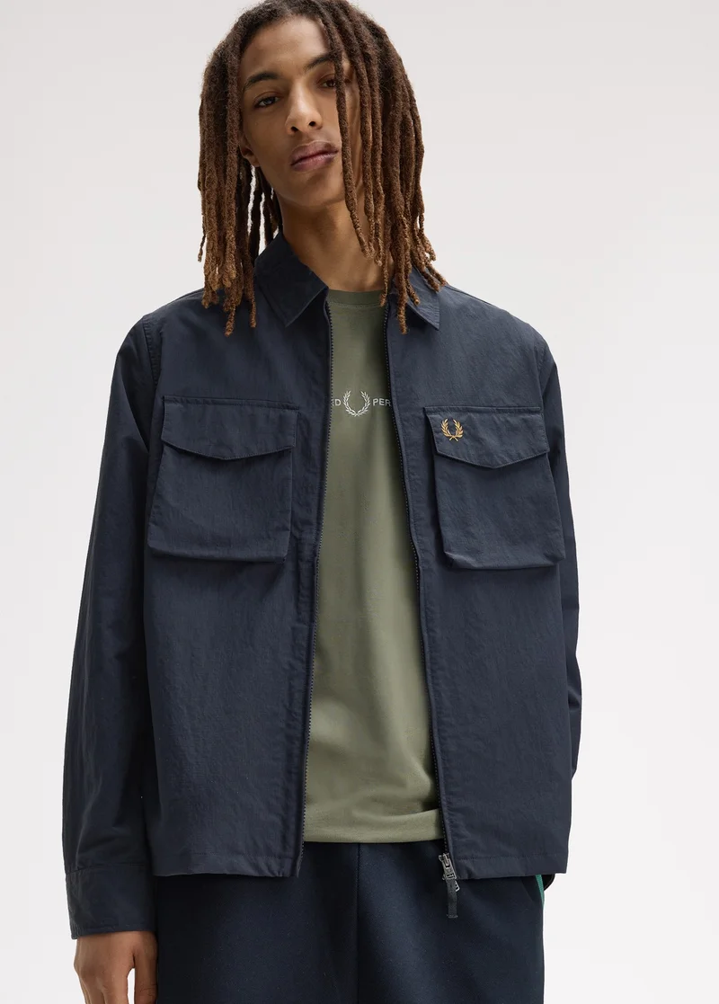 Layering: male model with shoulder-length locks looks into the camera and wears a green T-shirt layered with a navy double flap pocket overshirt in a crinkle textured material