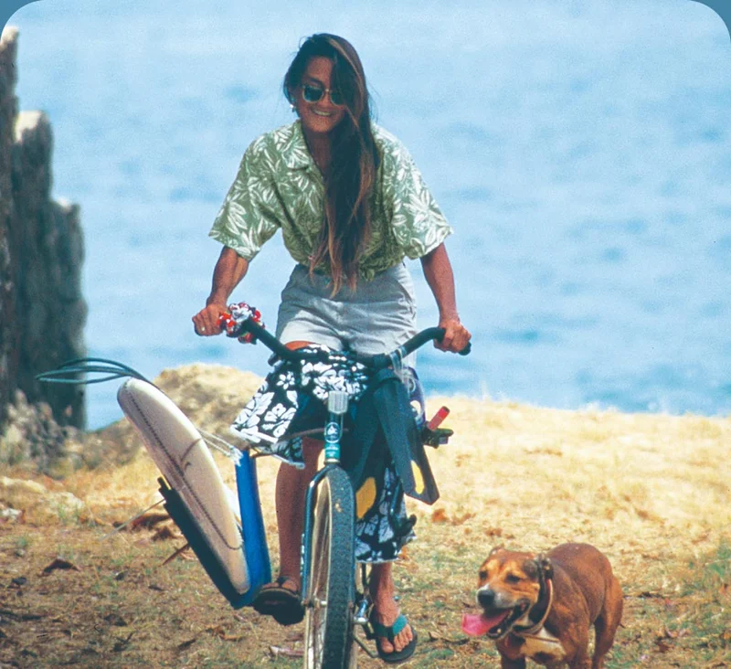 A person leaving the beach on their bike with a surfboard in the rack and a dog running beside.