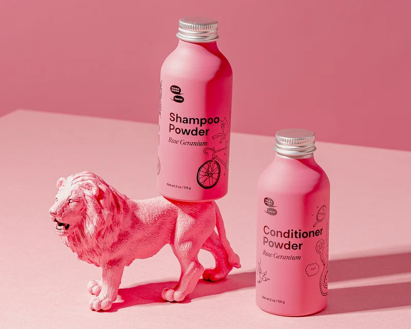 Two pink bottles of haircare products stand against a yellow backdrop with carrots coming out of the ground.