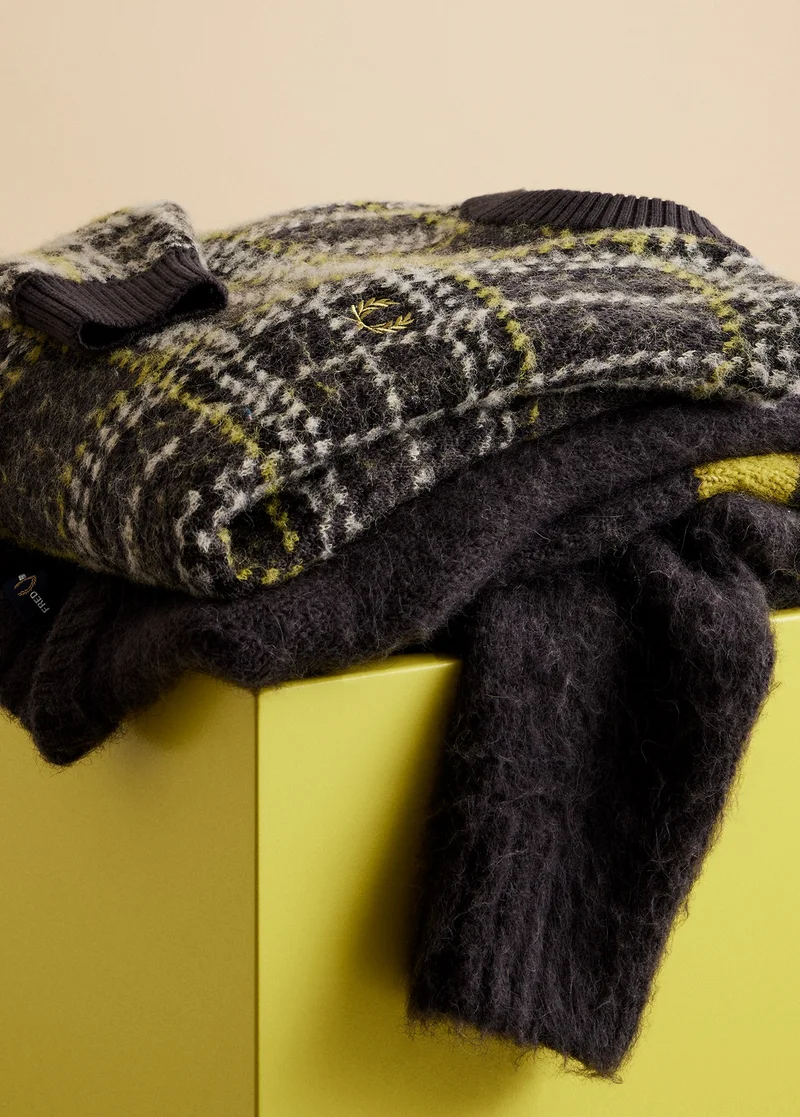 Image is of a folded up dark grey knitted jumper with tennis yellow and white chequered stitching, sitting on top of another dark grey knitted jumper with horizontal tennis yellow stripes on the upper sleeves. Both jumpers are placed casually on a tennis yellow coloured plinth, with one of the arms draped over the side.