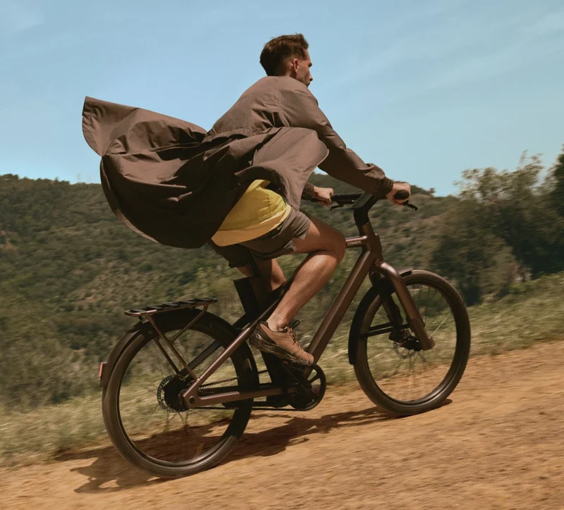 A photograph. A cyclist is riding a Cowboy bike over rugged, hilly terrain, his coat billows out behind him.