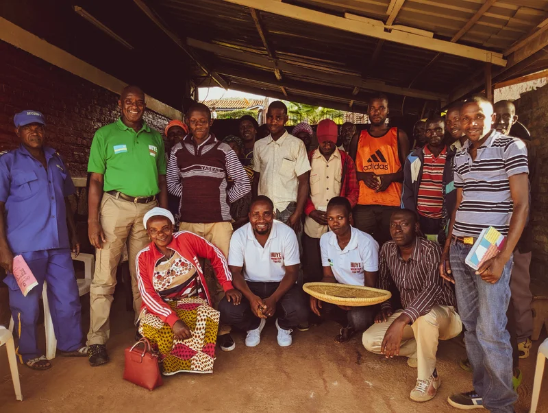 Group portrait of coffee producer David Rubanganzabo with Huye Mountain staff and local producers gathered under a covered workspace, smiling together; several wear Huye Mountain Coffee shirts, and one person holds a woven coffee sorting tray.