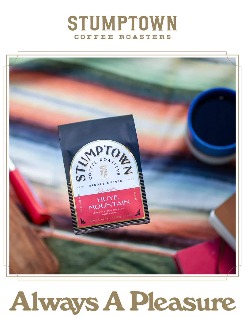Overhead view of a Stumptown Coffee Roasters “Huye Mountain” Rwanda single origin coffee bag on a marble countertop, with a small cup of black coffee to the right and a wooden cutting board in the background. Text below reads “Always A Pleasure.”