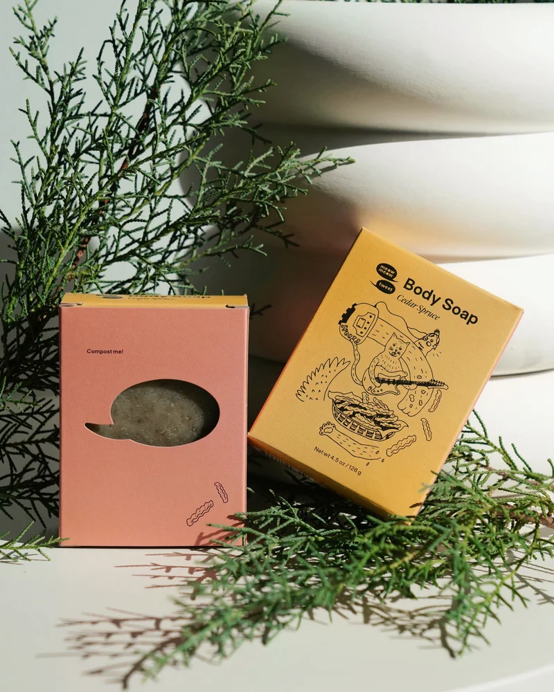 A mustard yellow box leans against sprigs of juniper and a ribbed vase
