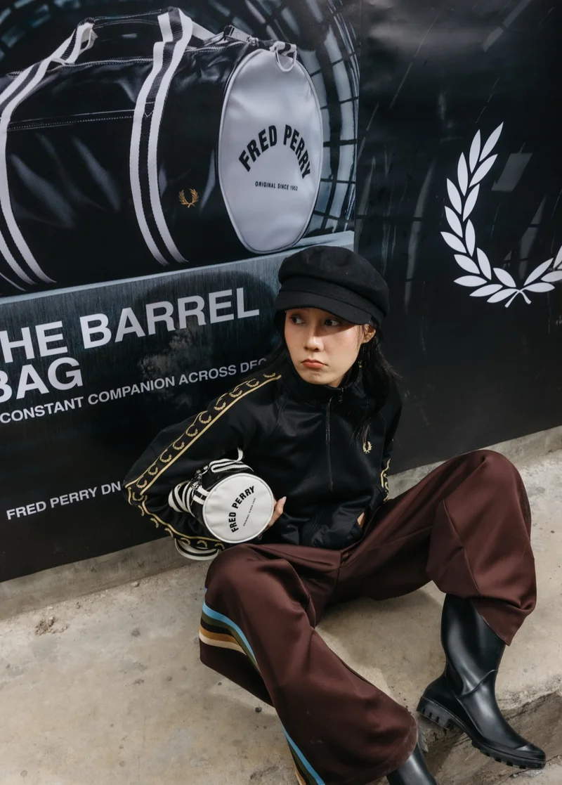 Female model sits on the side of the road on a walkway in front of a large Fred Perry barrel bag poster and wears the black and champagne track jacket while holding a mini barrel bag under her arm