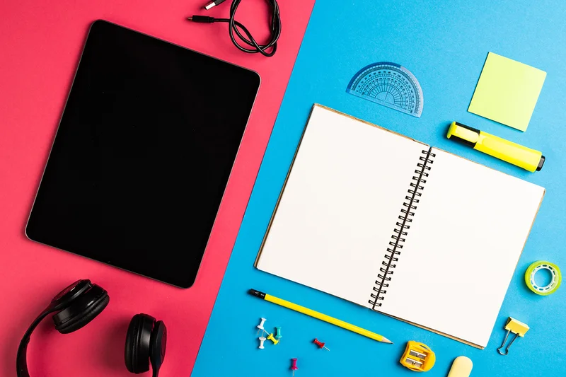 An image of a tablet and a notebook on a pink and blue surface surrounded by office supplies.