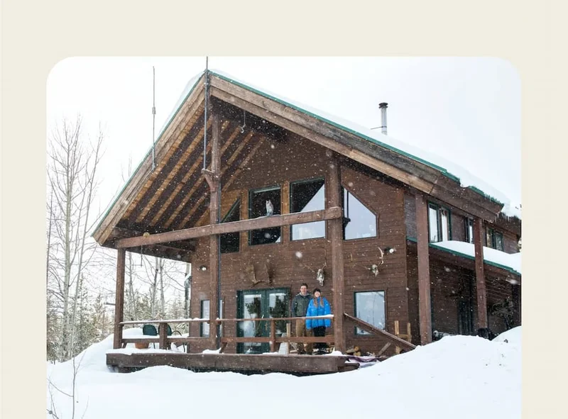Two people stand on the porch of a cabin on a snowy day.