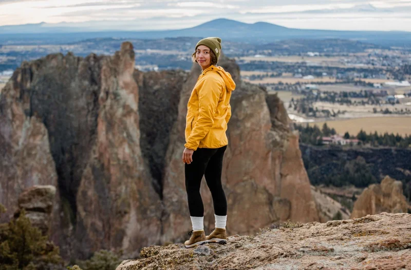 A young woman wearing a bright yellow jacket and a beige beanie stands on a rocky ledge overlooking a cliff. In the background, a vast landscape stretches out featuring distant mountains and a sprawling valley dotted with fields and buildings.