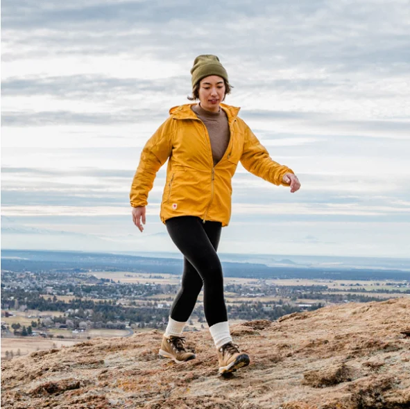A young woman in a bright yellow jacket and beige beanie is walking down a rocky hill with a panoramic view of a wide valley and distant mountains in the background.