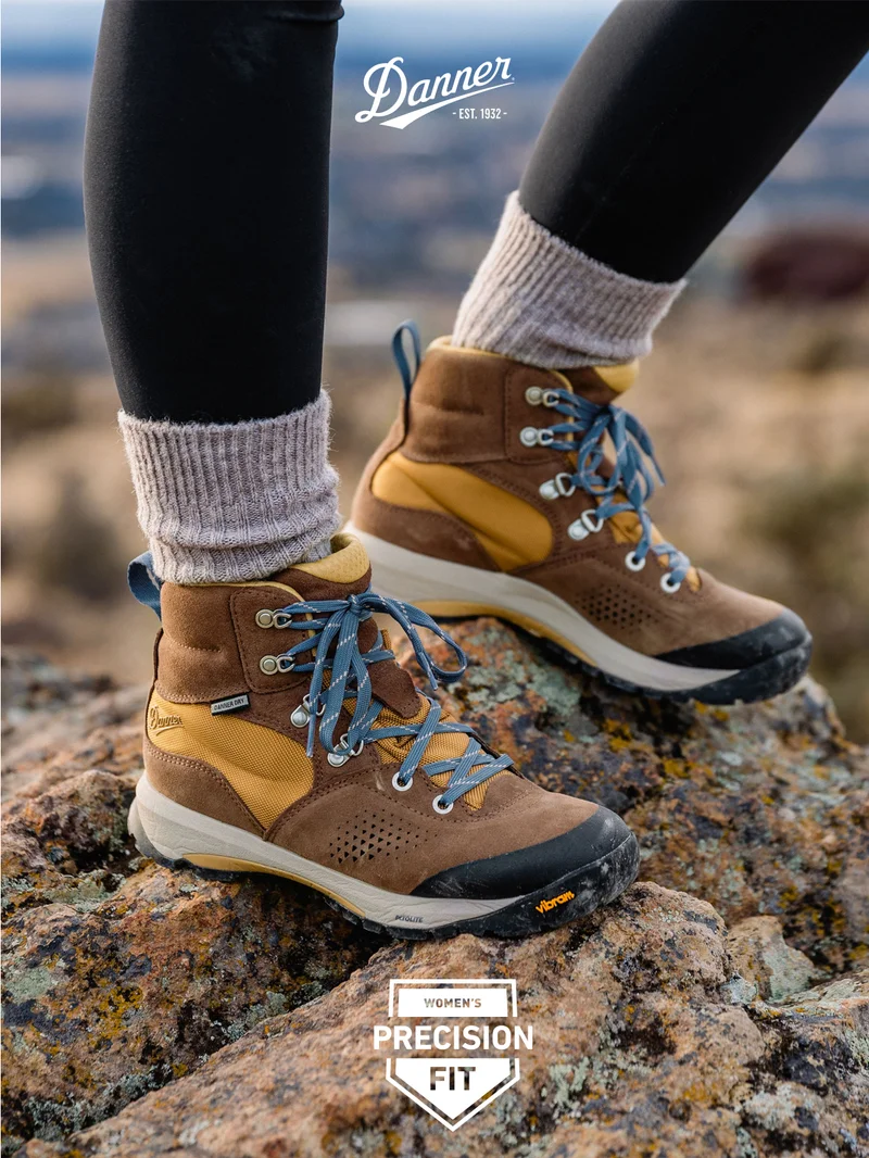 Close-up view of a woman's legs wearing Danner Women's Inquire Mid boots in Pinecone Brown/Prairie Sand on rocky terrain. The boots are detailed with blue laces and feature a durable Vibram outsole. The Danner logo is prominently displayed at the top, with the text "Women's Precision Fit" in a badge at the bottom.