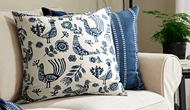 The KRUTBRÄNNARE cushion cover has an off-white background with a vibrant, rustic design of blue flowers, leaves and birds.
