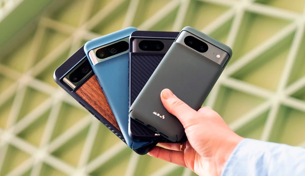 A hand holds four phones fanned out: One has a wood detailing case. The second has a blue case. The third has a dark blue case with angled lines etched in it. The last has a gray case with a small logo at the bottom that reads “Mous.”