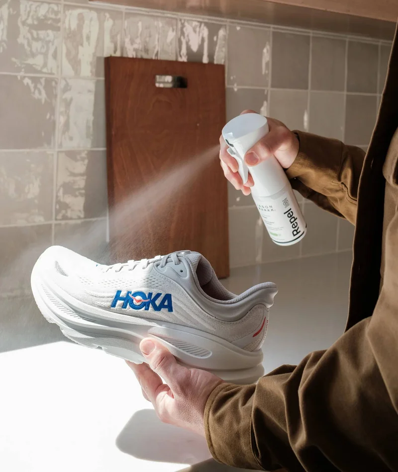 A medium shot captures a person's hands holding a white HOKA running shoe while spraying it with a white bottle labeled 'Repel' and 'JASON MARKK.'. Th...