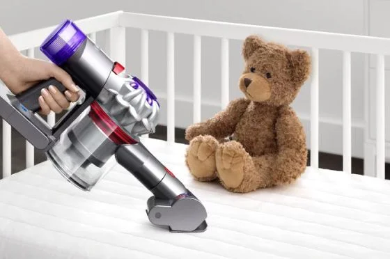 A Dyson Car+Boat vacuum with the Mini motorized head cleans a baby’s cot mattress with a teddy bear in the background.