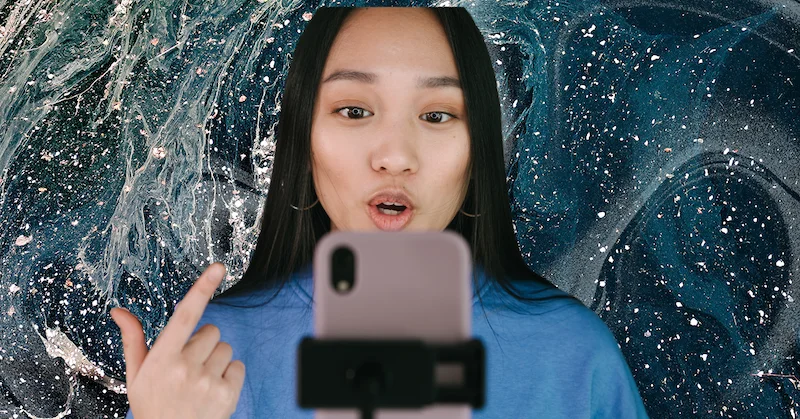 A young woman in a blue sweater records a video of herself on her phone on a blue-and-white swirl background.