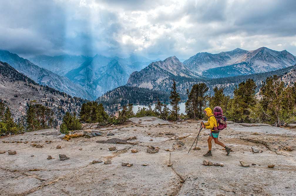 A hiker walks a trail high in the mountains with a storm brewing in the sky.