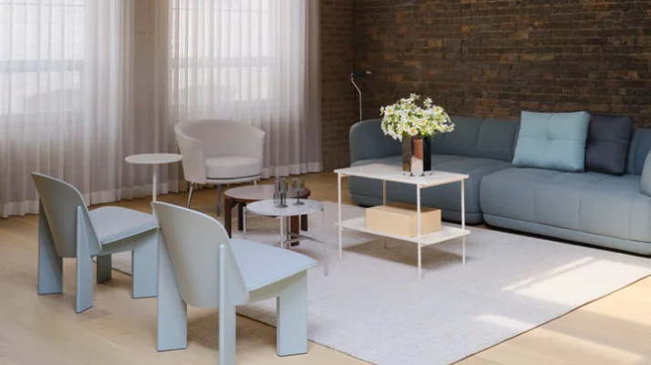 A lounge setting with windows on the left wall and a brick back wall behind the gray Quilton Sofa that faces the Chisel Lounge Chairs in pale blue, a Dorso Lounge Chair in white, a Rey Coffee Table, and a Rebar Side and Coffee Table.