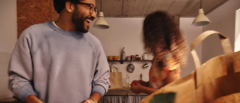 A smiling man prepares food in a kitchen with a woman in the background. Kitchen utensils hang on the back wall.