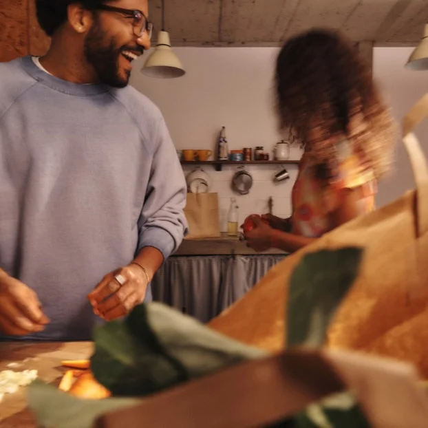 A smiling man prepares food in a kitchen with a woman in the background. Kitchen utensils hang on the back wall.