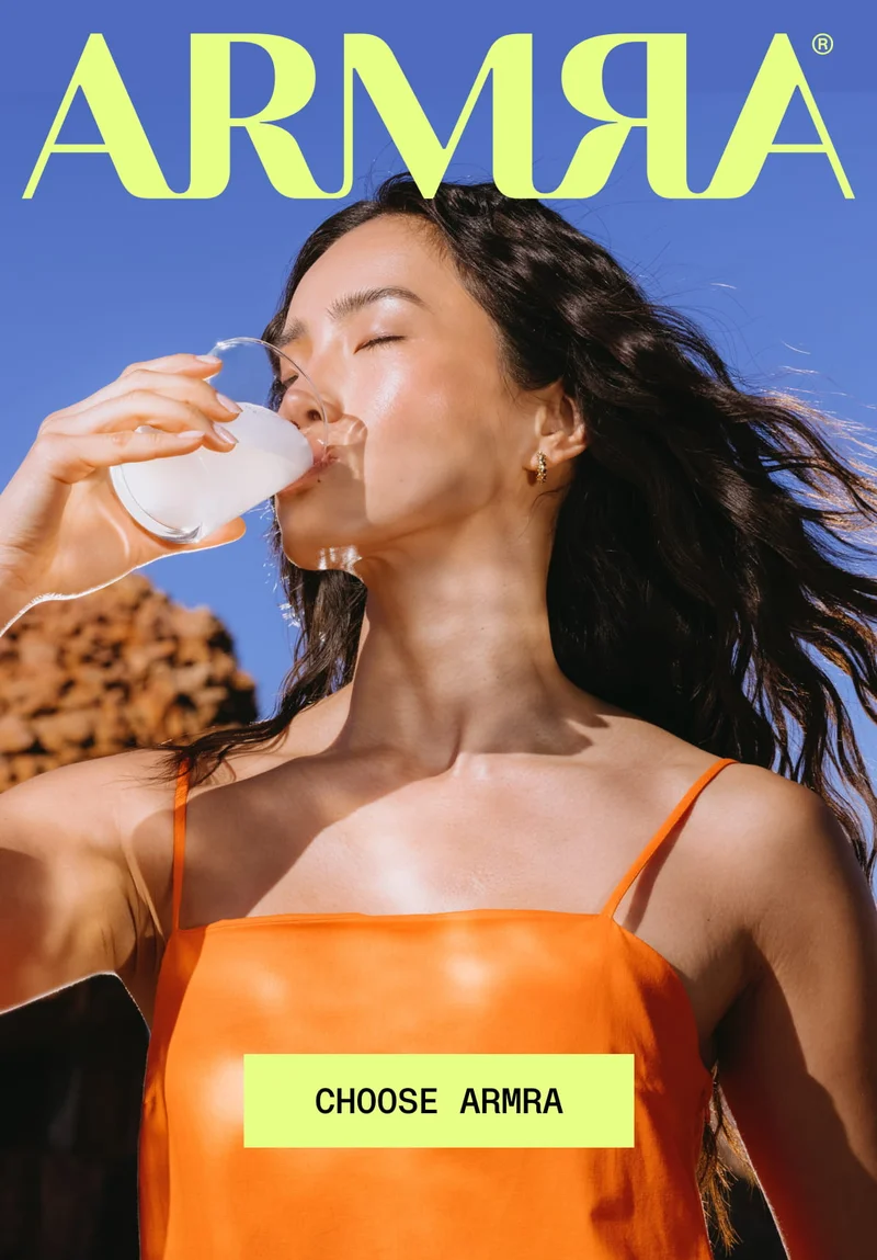 ARMRΑ® CHOOSE ARMRΑ. Woman in orange dress drinking a white ARMRΑ beverage from a glass outdoors in bright sunlight, blue sky and rocky desert background.