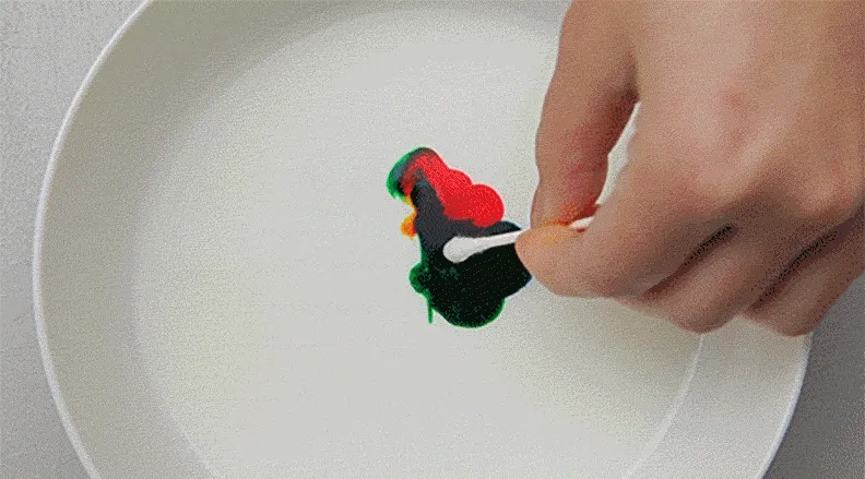 An overhead view of a plate of milk with food coloring swirling dramatically as soap is added. A child’s hand is visible in action.
