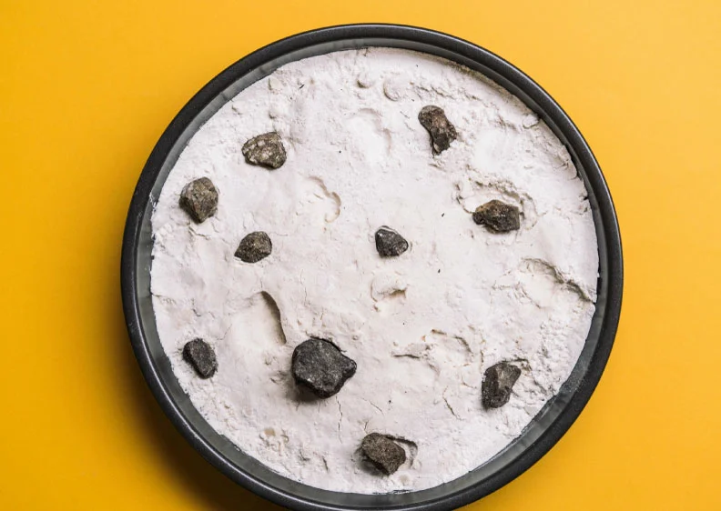 A round black tray filled with white flour shows rock-like objects scattered across it, mimicking the moon’s cratered surface. Set against a vivid yellow background.