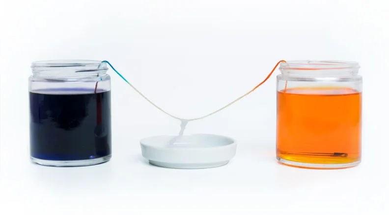 Two jars, one with blue liquid and one with orange, connected by a string over small white dishes, represent a salt crystal growth experiment.