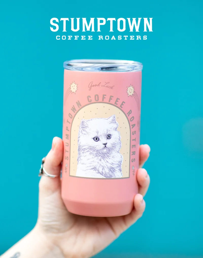 Hand holding a pink Stumptown Coffee Roasters tumbler featuring an illustrated white cat and “Good Luck” text against a bright teal background.
