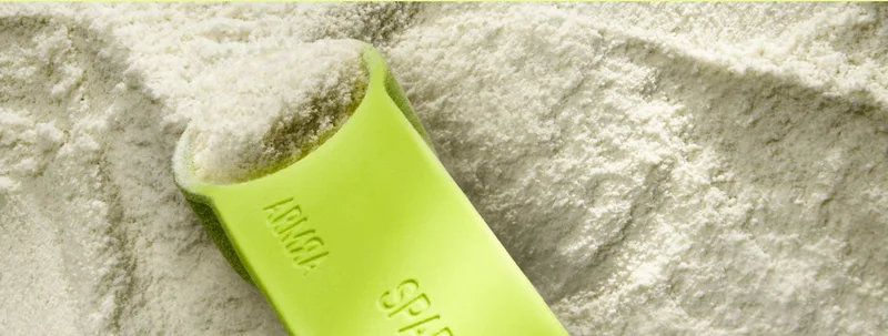 Close-up of a green scoop filled with ARMRA Colostrum powder resting in a bed of fine white powder.