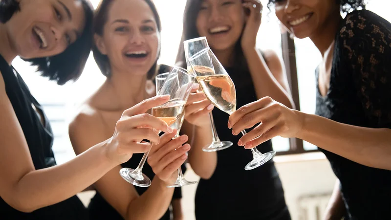 Four women wearing black cocktail dresses and clinking champagne glasses