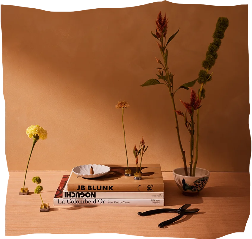 An artistic setup with dried flowers and decor items on a wooden desk against a peach backdrop, highlighting 'The Room by Goodee' event at Ace Hotel from March 12-19.