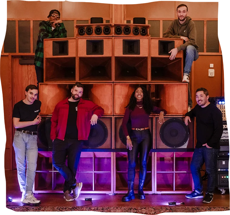 A group of six people pose casually in front of a large, vintage sound system with a vibrant energy that suggests a lively and immersive dub music experience.