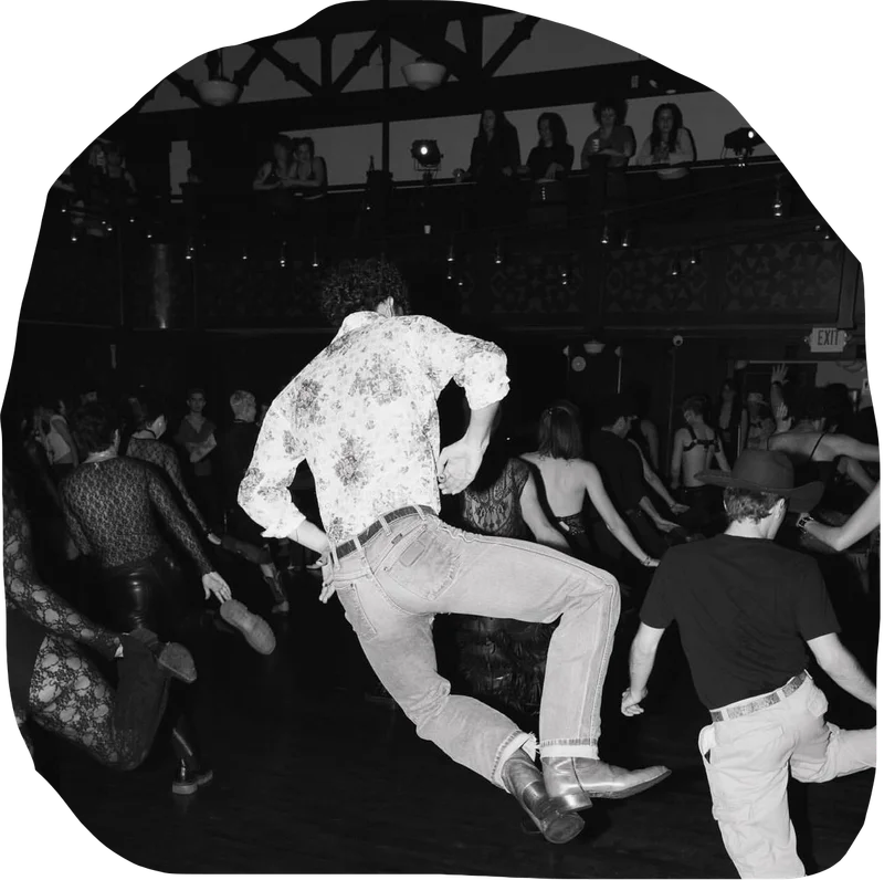 A black and white photo showing a man in Western attire energetically jumping and clicking his heels amidst a crowd of people dancing at the 'Stud Country Tea Dance'