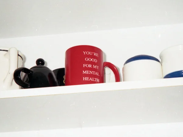 image of the We're Not Really Strangers You're Good For My Mental Health red mug on a shelf with a black ceramic tea pot ands some white mugs