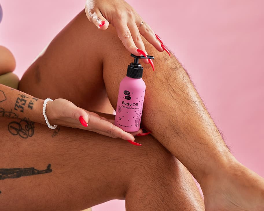 A pink bottle is held by a red nailed hand against hairy legs