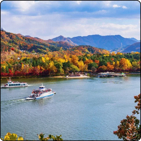Image Description: Boats on the water with mountains and trees with Fall-colored foliage in the background.