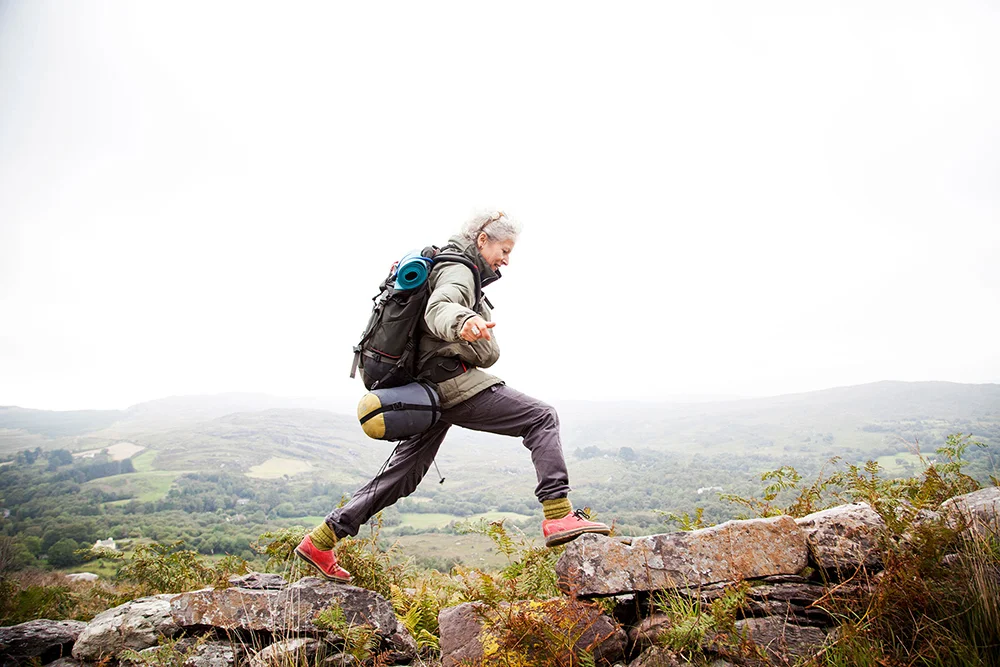 Older woman, with a backpacking back pack, a light gray coat, and dark gray pants, happily trekking in the mountains of Ireland.