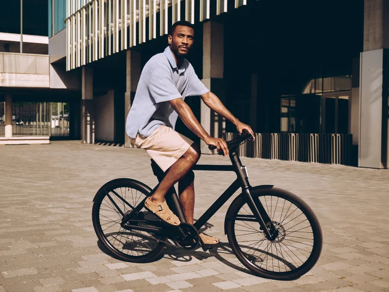 A man riding a black Cowboy e-bike in an urban setting, with modern architecture and a paved courtyard in the background.