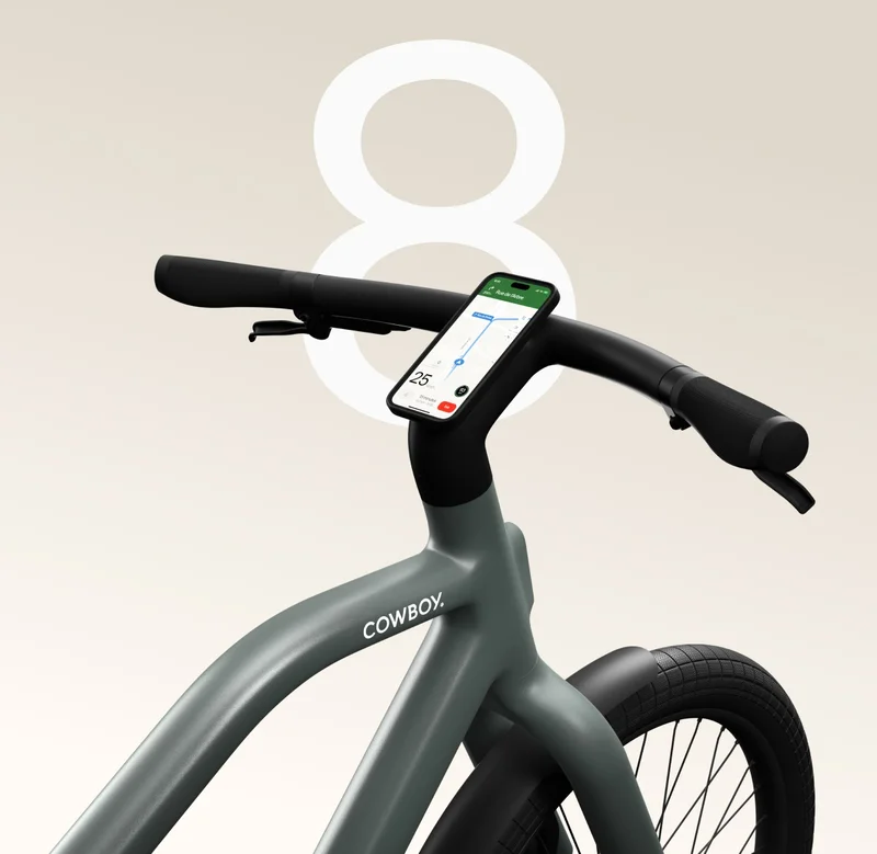 Close-up of the handlebars of a green Cowboy e-bike, with a smartphone mounted on the stem displaying navigation on the screen, and a large number “8” in the background.