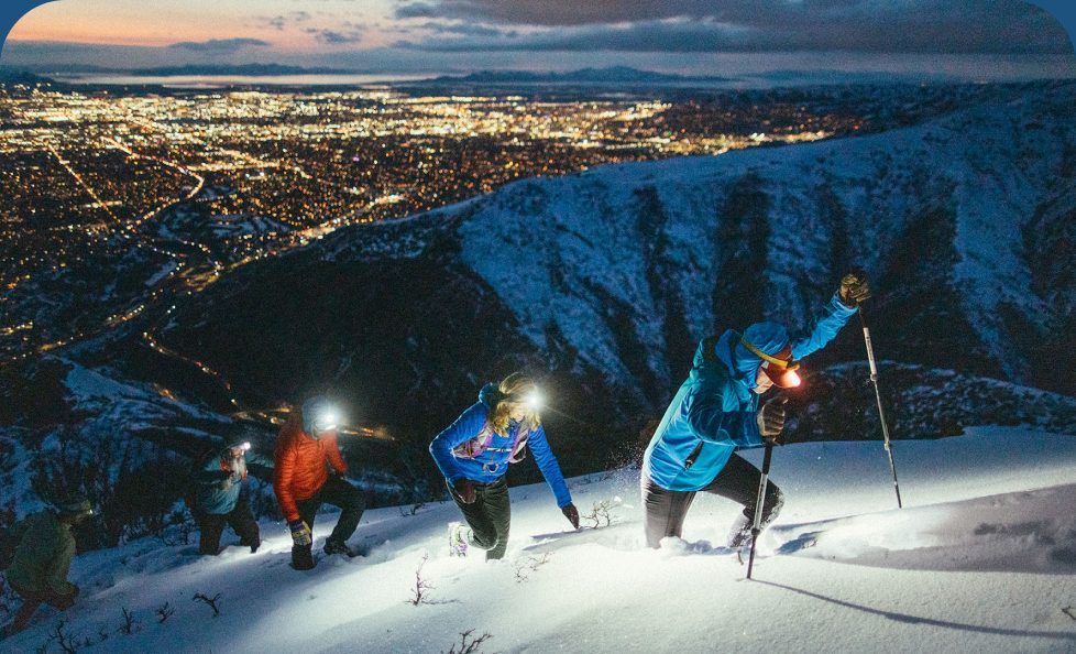 Runners break trail up a snowy mountain at night with city lights behind them.