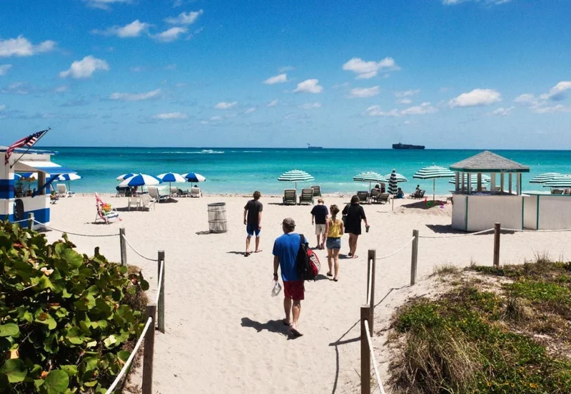 Image Description: Beachgoers strolling on the white sands of Miami Beach, surrounded by green shrubs and clear blue waters