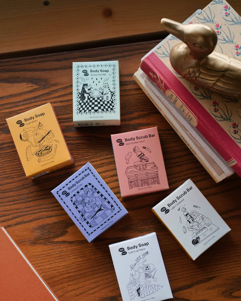 Six bars of soap sit on a wooden table next to a stack of books and a bronze duck